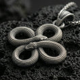 Load image into Gallery viewer, Oxidized snake knot pendant with intertwined serpents, detailed scale texture, and symmetrical looped design displayed on dark volcanic rock in a close-up luxury jewelry composition