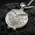 Load image into Gallery viewer, Polished silver owl necklace with intricate feather texture, circular medallion form, and expressive wise bird design placed on textured volcanic rock in a premium studio product shot