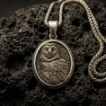 Load image into Gallery viewer, Bronze owl pendant with detailed feather engraving, oval medallion frame, and calm nocturnal bird silhouette displayed on dark volcanic rock in a close-up luxury jewelry composition