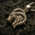 Load image into Gallery viewer, Bronze Aries ram pendant with detailed horn curve, textured goat head relief, and zodiac-inspired animal silhouette displayed on dark volcanic rock in a close-up luxury jewelry composition