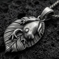 Load image into Gallery viewer, Oxidized frog pendant with smooth amphibian relief, leaf-shaped base, and nature-inspired animal silhouette displayed on dark volcanic rock in a close-up luxury jewelry composition