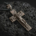 Load image into Gallery viewer, Bronze symbolic cross pendant with an oxidized finish resting on textured volcanic rock, captured in a close-up premium studio composition.