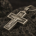 Load image into Gallery viewer, Bronze Cristo Protetor cross pendant necklace with an oxidized finish resting on textured volcanic rock, captured in a close-up premium studio composition.