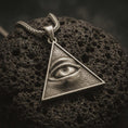 Load image into Gallery viewer, Bronze all-seeing eye pendant necklace with an oxidized finish resting on textured volcanic rock, captured in a close-up premium studio composition.