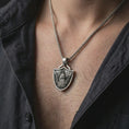 Load image into Gallery viewer, Oxidized Fortiores Una eagle shield pendant necklace worn on the neck, styled in a dark lifestyle portrait with an open shirt and masculine presentation.
