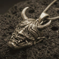 Load image into Gallery viewer, Bronze Oni mask pendant necklace with an oxidized finish resting on textured volcanic rock, captured in a close-up premium studio composition.