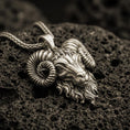 Load image into Gallery viewer, Bronze ram head pendant necklace with an oxidized finish resting on textured volcanic rock, captured in a close-up premium studio composition.
