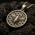 Load image into Gallery viewer, Bronze bee pendant necklace with an oxidized finish resting on textured volcanic rock, captured in a close-up premium studio composition.