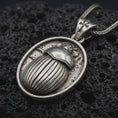 Load image into Gallery viewer, Sterling silver Egyptian scarab pendant necklace with an oxidized finish resting on textured volcanic rock, captured in a close-up premium studio composition.