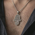 Load image into Gallery viewer, Oxidized Jerusalem Hamsa hand pendant necklace worn on the neck, styled in a dark lifestyle portrait with an open shirt and meaningful presentation.