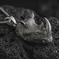 Load image into Gallery viewer, Sterling silver rhinoceros head pendant necklace with an oxidized finish resting on textured volcanic rock, captured in a close-up premium studio composition.