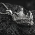 Load image into Gallery viewer, Polished silver rhinoceros head pendant necklace displayed on black volcanic rock, shown in a refined macro product shot with lifelike horn detail, textured skin relief, and bold wildlife sculpture design.