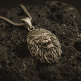Load image into Gallery viewer, Bronze roaring lion head pendant necklace with an oxidized finish resting on textured volcanic rock, captured in a close-up premium studio composition.