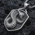 Load image into Gallery viewer, Sterling silver cobra shield pendant necklace with an oxidized finish resting on textured volcanic rock, captured in a close-up premium studio composition.