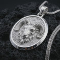Load image into Gallery viewer, Polished silver ram head pendant necklace displayed on black volcanic rock, shown in a refined macro product shot with curled horns, sculpted facial detail, and oval medallion frame.