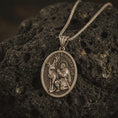 Load image into Gallery viewer, Bronze Saint Hubert pendant necklace with an oxidized finish resting on textured volcanic rock, captured in a close-up premium studio composition.
