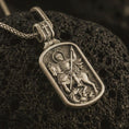 Load image into Gallery viewer, Bronze Saint George dog tag pendant necklace with an oxidized finish resting on textured volcanic rock, captured in a close-up premium studio composition.