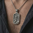 Load image into Gallery viewer, Oxidized Saint Benedict dog tag pendant necklace worn on the neck, styled in a dark lifestyle portrait with an open shirt and masculine presentation.