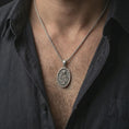 Load image into Gallery viewer, Oxidized Saint Rita pendant necklace worn on the neck, styled in a dark lifestyle portrait with an open shirt and masculine presentation.