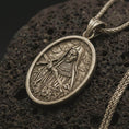 Load image into Gallery viewer, Bronze Saint Rita pendant necklace with an oxidized finish resting on textured volcanic rock, captured in a close-up premium studio composition.