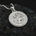 Load image into Gallery viewer, Polished silver Saint Benedict medal pendant necklace displayed on black volcanic rock, shown in a refined macro product shot with detailed saint figure and engraved Latin lettering.