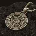 Load image into Gallery viewer, Bronze Saint Benedict medal pendant necklace with an oxidized finish resting on textured volcanic rock, captured in a close-up premium studio composition.