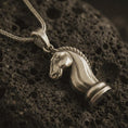 Load image into Gallery viewer, Bronze knight chess piece pendant necklace with an oxidized finish resting on textured volcanic rock, captured in a close-up premium studio composition.