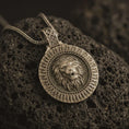 Load image into Gallery viewer, Bronze Viking lion medallion pendant necklace with an oxidized finish resting on textured volcanic rock, captured in a close-up premium studio composition.