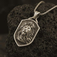 Load image into Gallery viewer, Bronze lion shield pendant necklace with an oxidized finish resting on textured volcanic rock, captured in a close-up premium studio composition.