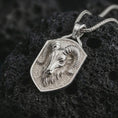 Load image into Gallery viewer, Ram head shield pendant necklace in polished silver resting on dark volcanic rock, captured in a close-up studio image that highlights the sculpted Aries-inspired face and horn detail.