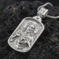 Load image into Gallery viewer, Polished silver Sacred Heart pendant necklace displayed on black volcanic rock, shown in a refined macro product shot with detailed Christ figure and engraved background pattern.