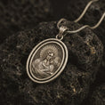 Load image into Gallery viewer, Bronze Madonna and Child pendant necklace resting on black volcanic stone, featuring an oval double-frame medallion with the Virgin Mary holding baby Jesus in a detailed devotional relief.