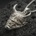 Load image into Gallery viewer, Polished silver Oni mask pendant necklace displayed on black volcanic rock, shown in a refined macro product shot with detailed horns, fangs, and engraved Japanese-inspired mask relief.