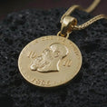 Load image into Gallery viewer, Gold plated heraldic eagle coin pendant necklace photographed on porous black volcanic stone, emphasizing the regal emblem, round medallion silhouette, and symbolic laurel accents in a luxury jewelry composition.