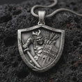 Load image into Gallery viewer, Sterling silver samurai shield pendant necklace with an oxidized finish resting on textured volcanic rock, captured in a close-up premium studio composition.