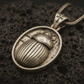 Load image into Gallery viewer, Bronze Egyptian scarab pendant necklace with an oxidized finish resting on textured volcanic rock, captured in a close-up premium studio composition.