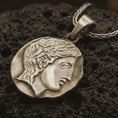 Load image into Gallery viewer, Bronze ancient Greek coin pendant necklace resting on black volcanic stone, featuring a classical female profile in an irregular coin-shaped relief with antique-inspired surface texture.