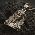 Load image into Gallery viewer, Bronze Tutankhamun pendant necklace with an oxidized finish resting on textured volcanic rock, captured in a close-up premium studio composition.