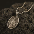 Load image into Gallery viewer, Bronze Saint Francis pendant necklace on textured volcanic rock, close-up studio shot highlighting the saint relief, bird, wolf detail, and antique silver contrast.