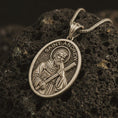 Load image into Gallery viewer, Bronze Saint Andrew pendant necklace on textured volcanic rock, close-up studio shot highlighting the apostle relief, cross, book detail, and antique silver contrast.