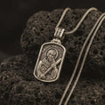 Load image into Gallery viewer, Bronze Saint Andrew dog tag pendant necklace on textured volcanic rock, close-up studio shot highlighting the apostle relief, cross, book detail, and antique silver contrast.