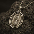 Load image into Gallery viewer, Bronze Our Lady of Guadalupe spinner pendant necklace on textured volcanic rock, close-up studio shot highlighting the Virgin Mary relief, radiant halo, crescent moon, and antique silver contrast.