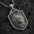 Load image into Gallery viewer, Wolf shield pendant necklace in oxidized sterling silver resting on black volcanic stone, shown in a detailed macro product photograph with textured fur, alert ears, and an angular shield frame.