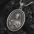 Load image into Gallery viewer, Oxidized Saint Francis pendant necklace on textured volcanic rock, close-up studio shot highlighting the saint relief, lamb, birds, and antique silver contrast.