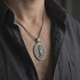 Load image into Gallery viewer, Oxidized Saint Michael spinning pendant necklace worn on the chest, close-up lifestyle jewelry photo showing the Christian protection medallion on a spiga chain with dark clothing.