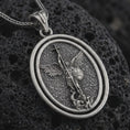 Load image into Gallery viewer, Oxidized Saint Michael spinning pendant necklace on textured volcanic rock, close-up studio shot highlighting the archangel wings, sword, shield, fallen demon, and antique silver contrast.