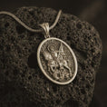 Load image into Gallery viewer, Bronze Saint George pendant necklace on textured volcanic rock, close-up studio shot highlighting the mounted saint, spear, dragon detail, and antique silver contrast.