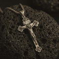 Load image into Gallery viewer, Bronze crucifix pendant necklace with an oxidized finish displayed on black volcanic stone, captured in a close-up studio image highlighting the detailed Jesus figure and floral cross tips.