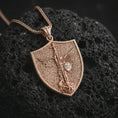 Load image into Gallery viewer, Rose gold plated Saint Michael pendant necklace photographed on porous volcanic stone, emphasizing the shield silhouette, sacred angel figure, and refined religious craftsmanship.