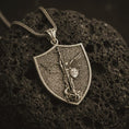 Load image into Gallery viewer, Bronze Saint Michael pendant necklace displayed on black volcanic stone, featuring a shield-shaped medal with Archangel Michael defeating evil in a detailed macro product photograph.